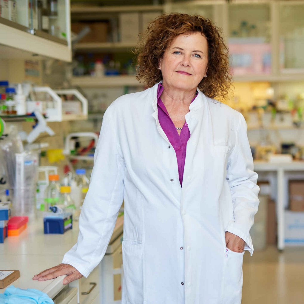 This is a picture of Erin Schuman, who has been director at the Max Planck Institute for Brain Research in Frankfurt since 2009. In this picture, Schuman is wearing a white lab coat buttoned over a purple shirt. She is standing in what appears to be a lab setting, with one hand resting on the counter, and the other one in the pocket of her lab coat. She is looking into the camera. Her brown hair is of medium lenght and curly.