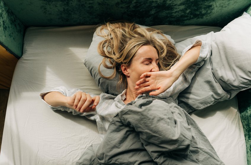 Nach der Zeitumstellung fällt es vielen Menschen schwer, in der Früh aufzustehen. Eine Frau streckt sich nach dem Aufwachen und gähnt im Bett liegend.