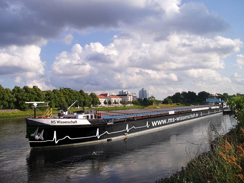 Das Frachtschiff MS Wissenschaft fährt entlang eines Flusses, vorbei an grünen Bäumen und städtischen Gebäuden unter einem bewölkten Himmel.