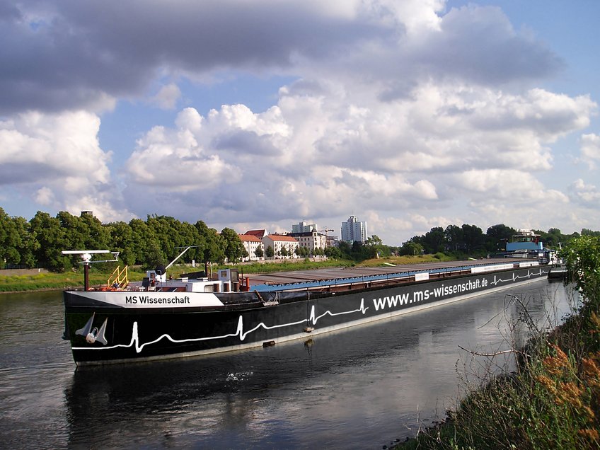 Am 7. Mai startet die MS Wissenschaft ihre neue Tour. Das Frachtschiff MS Wissenschaft fährt entlang eines Flusses, vorbei an grünen Bäumen und städtischen Gebäuden unter einem bewölkten Himmel.
