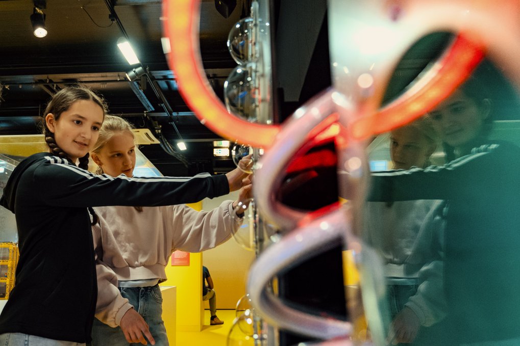 Zwei Schülerinnen experimentieren in einer wissenschaftlichen Ausstellung, umgeben von farbigen Lichtern und Röhren.