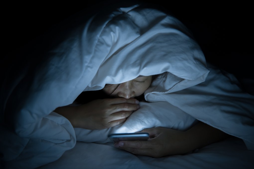 Person liegt im Dunkeln unter einer Decke und schaut auf ein Smartphone.