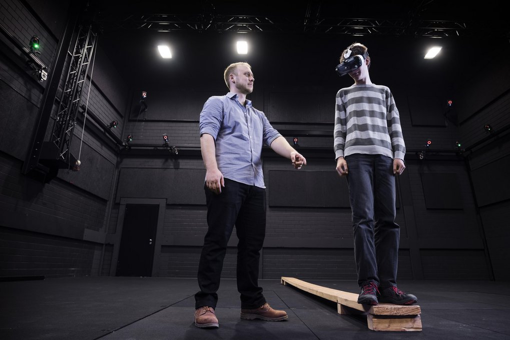 Two individuals interact in a dark room; one person is wearing a VR headset, standing on a wooden plank.