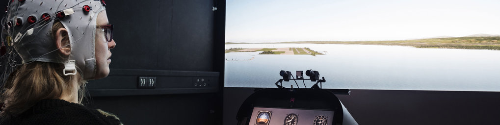 An individual wearing an EEG cap with sensors appears to be in a simulation room, observing a screen displaying a water landscape with distant land formations.