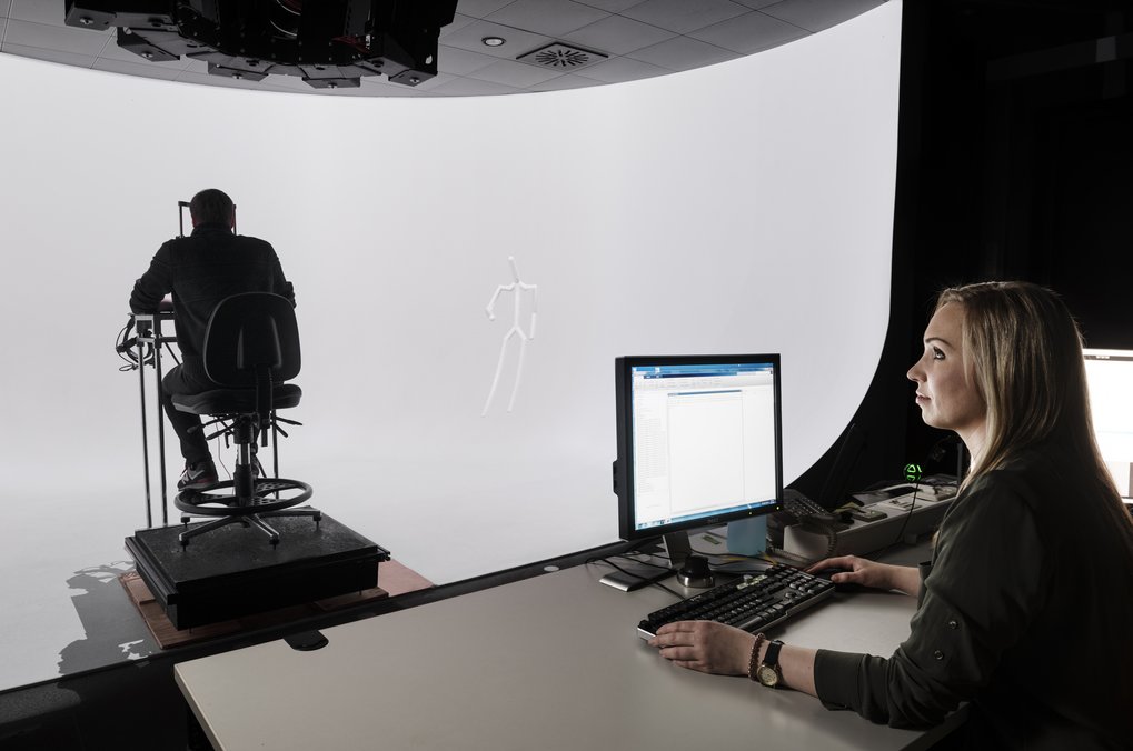 Two individuals in a studio setting, one at a computer desk, another seated on a platform facing a white screen.