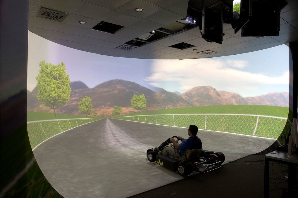 Individual driving a Go-Kart in a room with a curved screen showing a virtual countryside road.